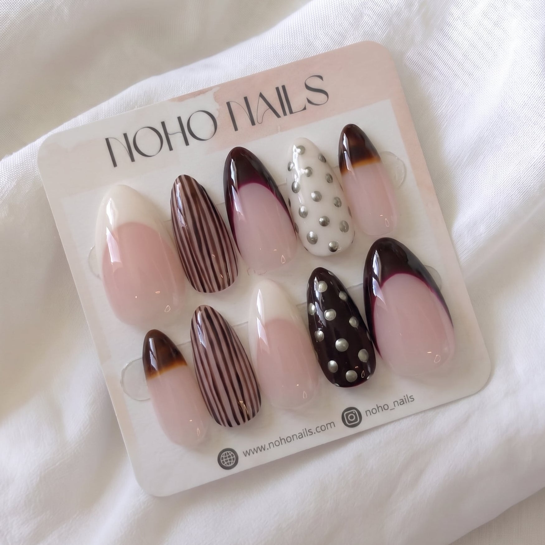 Noir et Crème press-on nails by Noho Nails: long almond nails with cream and black-cherry French tips, brown pinstripes, tortoiseshell tips and silver studs on a Noho Nails display card