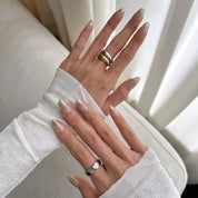 Hands wearing Golden Glaze press-on nails, sheer nude with speckled glitter and gold glitter tips, with gold and silver rings