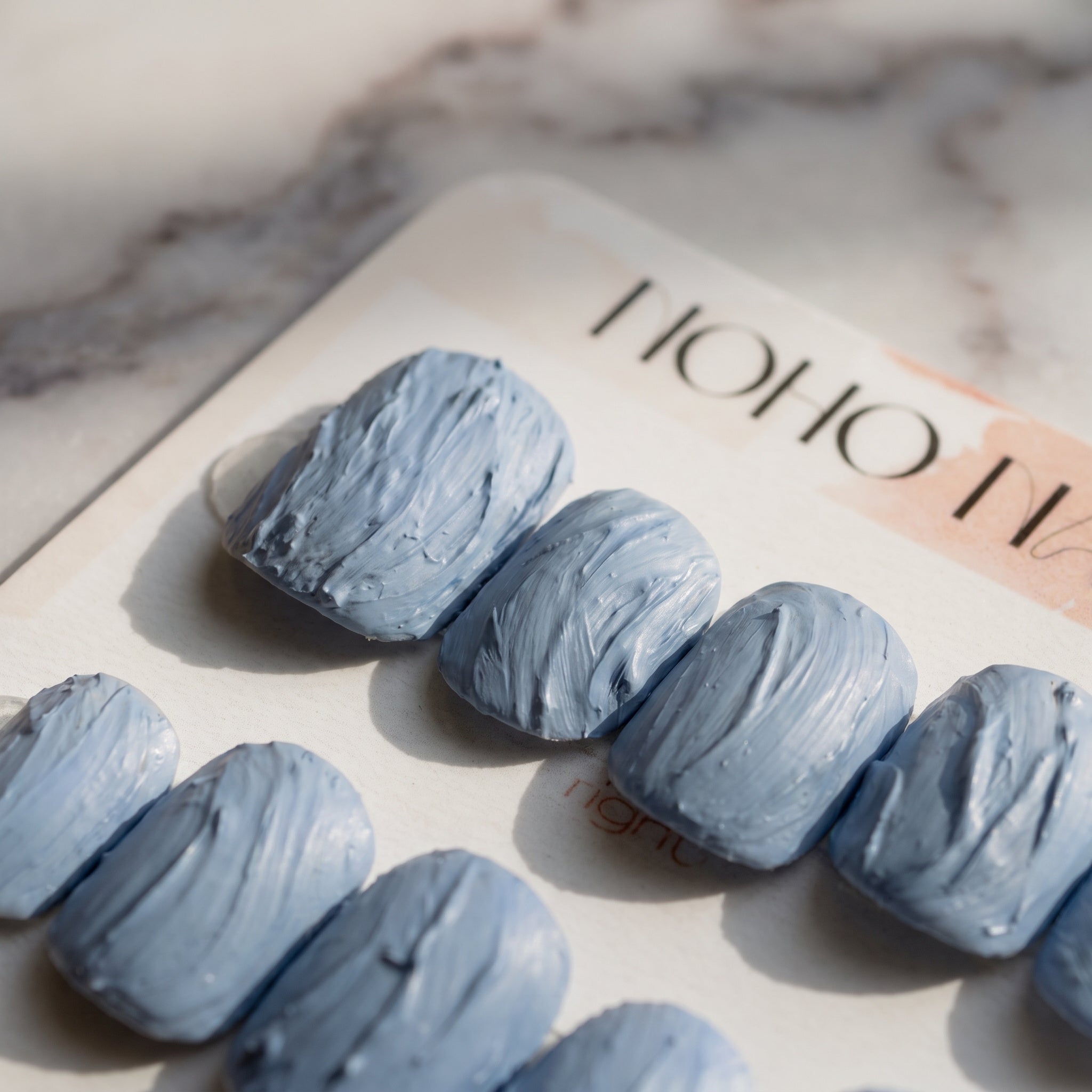 Angled close-up of Slate Relief press-on nails on a Noho Nails display card on white marble, showing the textured matte blue-gray finish