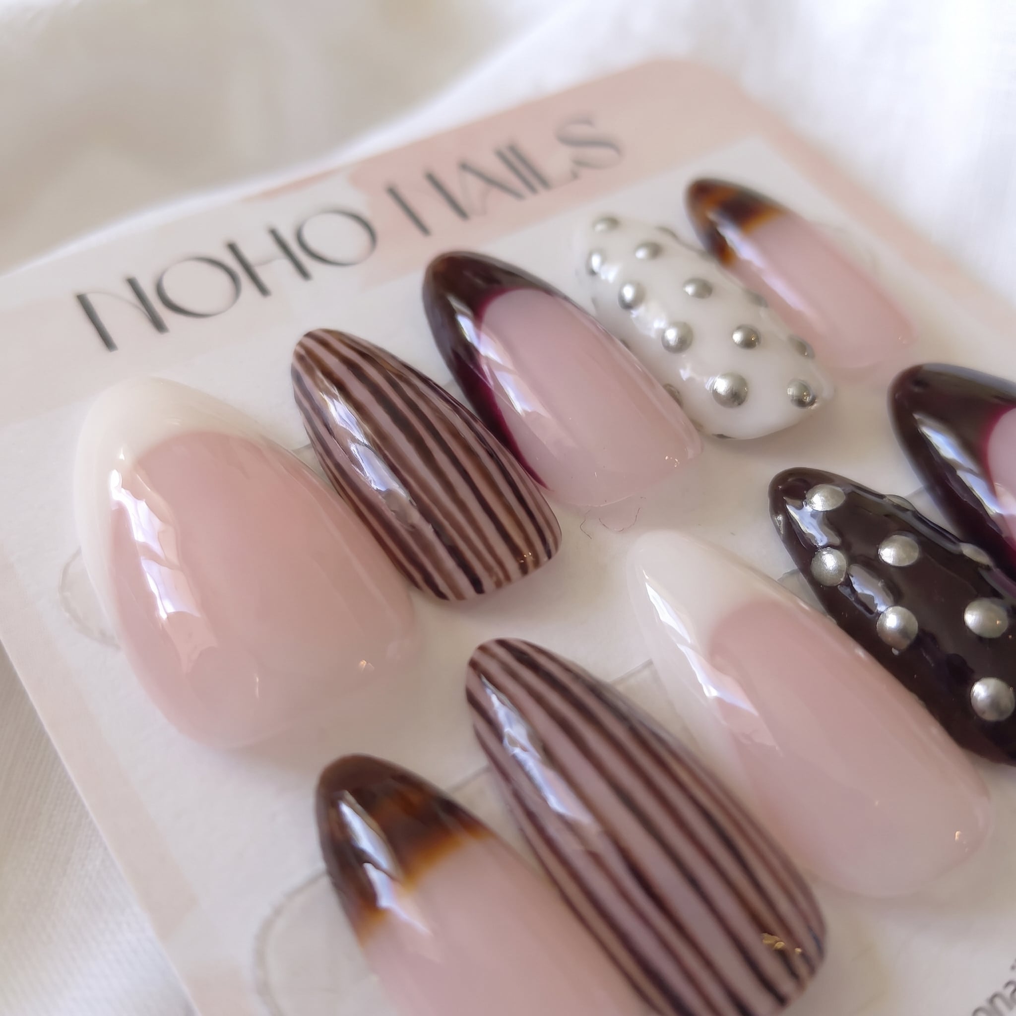 Close-up of Noir et Crème press-on nails by Noho Nails: long almond nails with cream and black-cherry French tips, brown pinstripes, tortoiseshell tips and silver studs