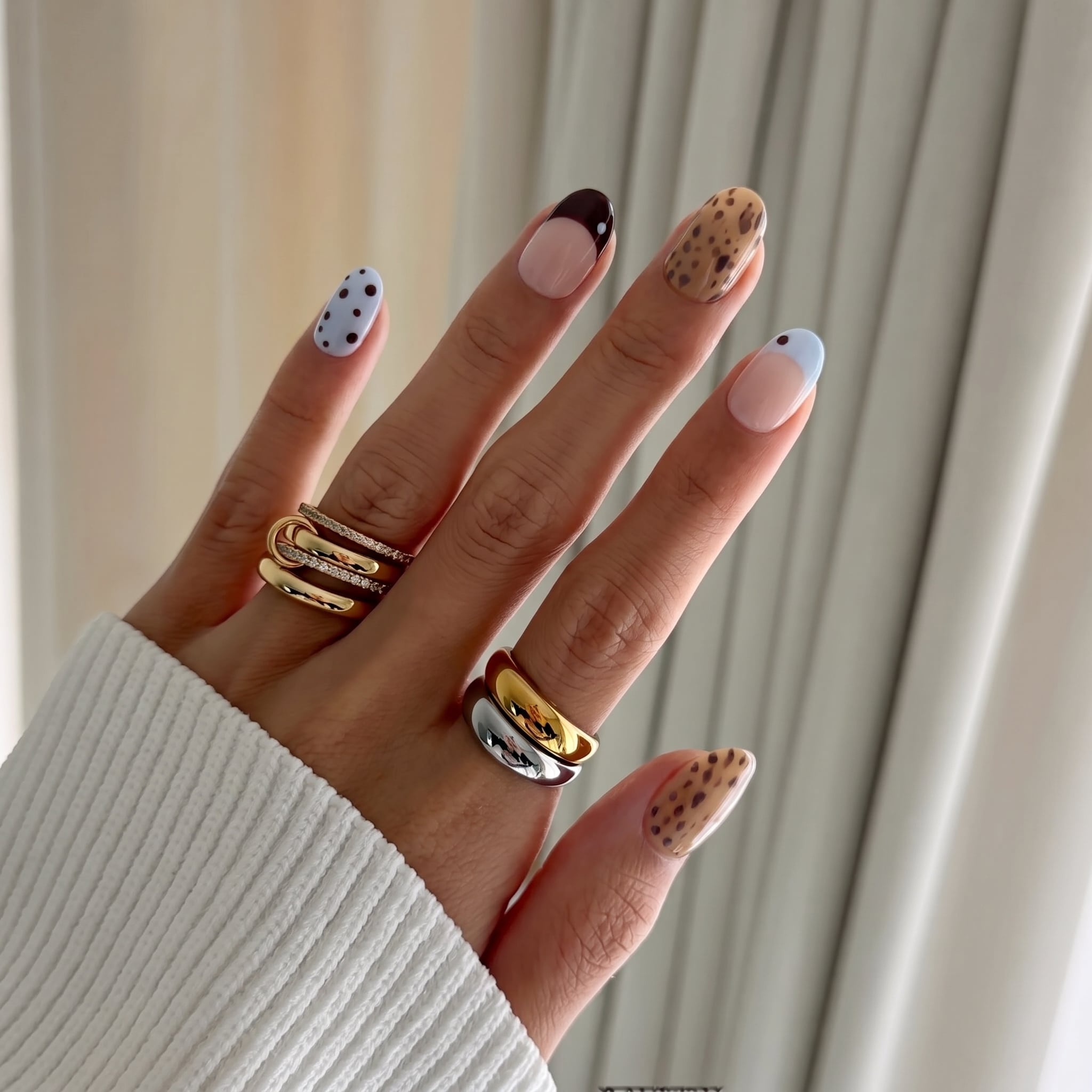 Hand wearing Petite Tache press-on nails: short round nails with caramel leopard spots, baby-blue polka dots and black-cherry and baby-blue French tips, with gold and silver rings
