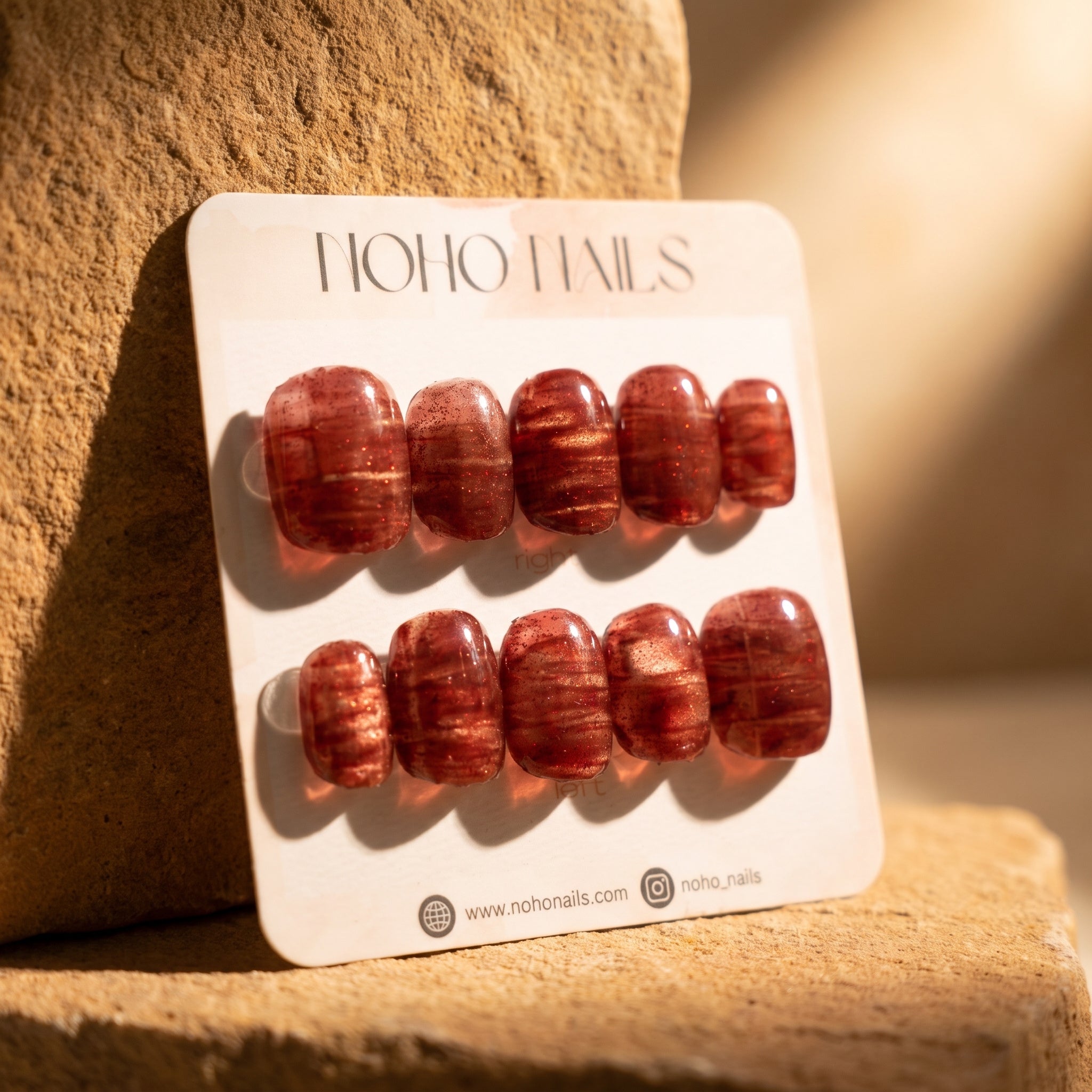 Cognac Emboss press-on nails on a Noho Nails display card propped against warm sandstone in golden light
