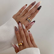 Hands wearing Noir et Crème press-on nails in a long almond shape, with cream and black-cherry French tips, brown pinstripes, tortoiseshell tips and silver-studded nails, and gold and silver rings