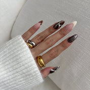 Hand wearing Café de Flore press-on nails in a long almond shape: dark brown cat-eye glitter with white line art, a tortoiseshell tip and a cream French tip, with gold and silver rings