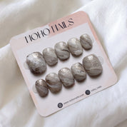 Sediment No. 9 press-on nails by Noho Nails: ten short squoval nails with hand-painted greige, taupe and chalky white swirls and gold flecks on a Noho Nails display card lying on white fabric