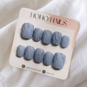 Slate Relief press-on nails by Noho Nails: ten short squoval nails in matte dusty blue-gray with a raised, hand-sculpted texture on a Noho Nails display card lying on white fabric