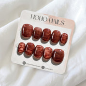 Cognac Emboss press-on nails by Noho Nails: ten short squoval nails in glossy deep cognac with a streaked copper texture on a Noho Nails display card lying on white fabric