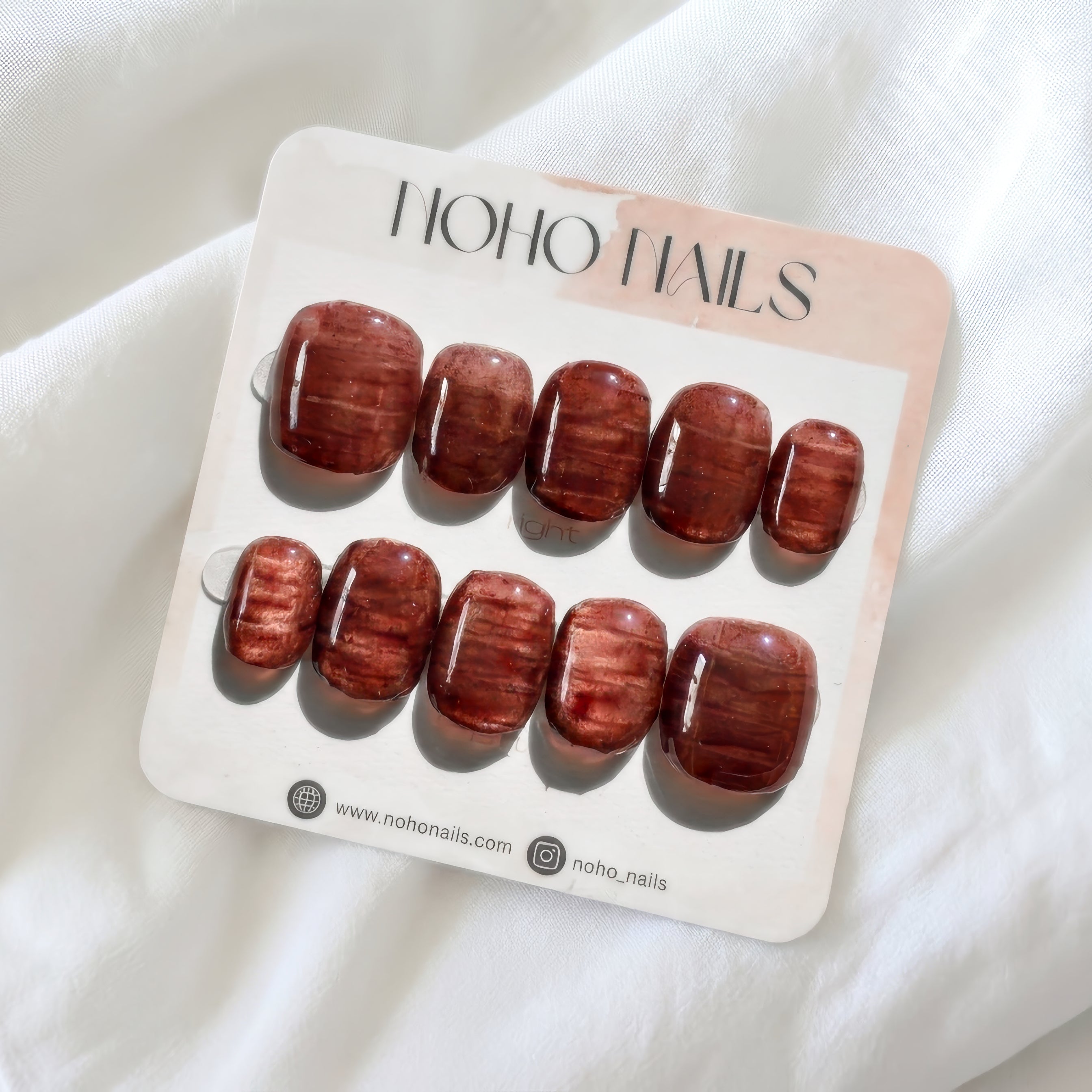 Cognac Emboss press-on nails by Noho Nails: ten short squoval nails in glossy deep cognac with a streaked copper texture on a Noho Nails display card lying on white fabric