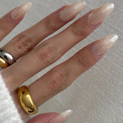 Hand wearing Prosecco Pearl press-on nails in a long almond shape, pale pink with champagne shimmer, with gold and silver rings