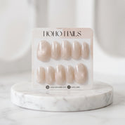 Prosecco Pearl press-on nail set on a Noho Nails card displayed on a marble stand, showing blush-pink nails with champagne cat-eye shimmer