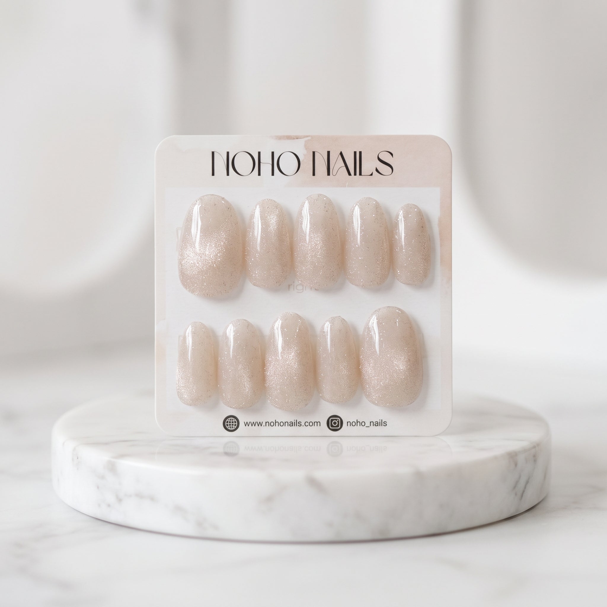 Prosecco Pearl press-on nail set on a Noho Nails card displayed on a marble stand, showing blush-pink nails with champagne cat-eye shimmer