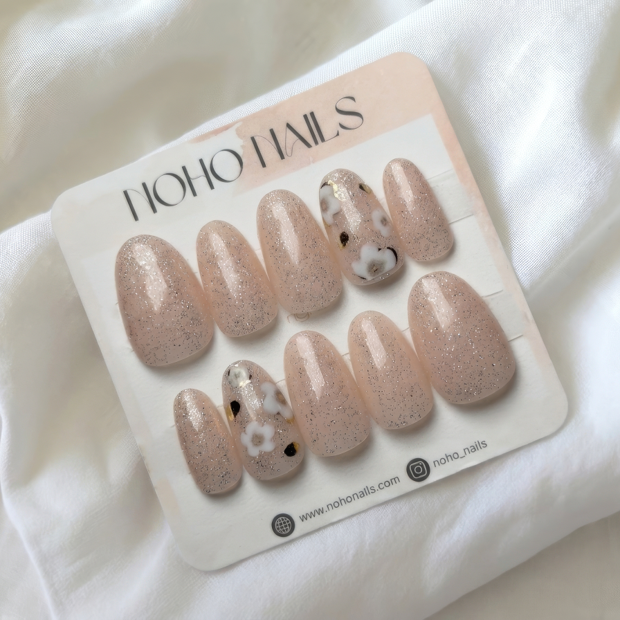 A set of press-on nails on a white card labeled HOHO NAILS, placed on a soft white fabric background. The nails are a sheer nude pink with fine glitter, and two feature a white base with black leopard spots.