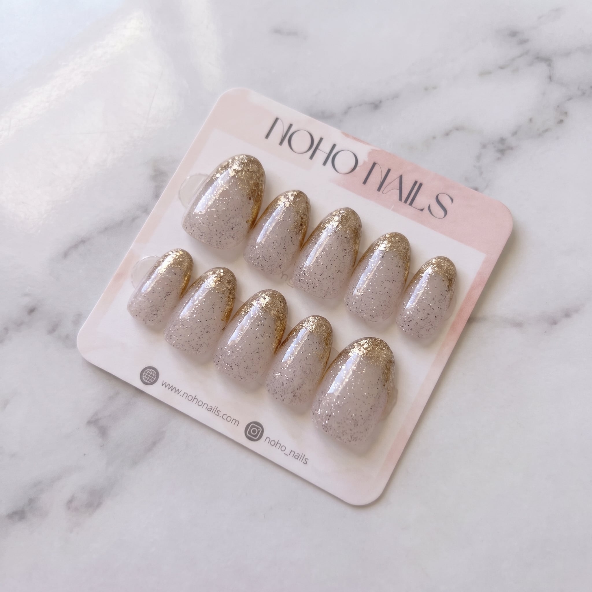 Golden Glaze press-on nails on a Noho Nails display card lying on white marble