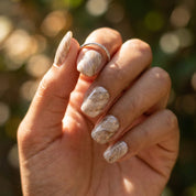 Hand wearing Sediment No. 9 press-on nails: greige, taupe and white marbled swirls with gold flecks, with a thin silver ring, against a sunlit green background