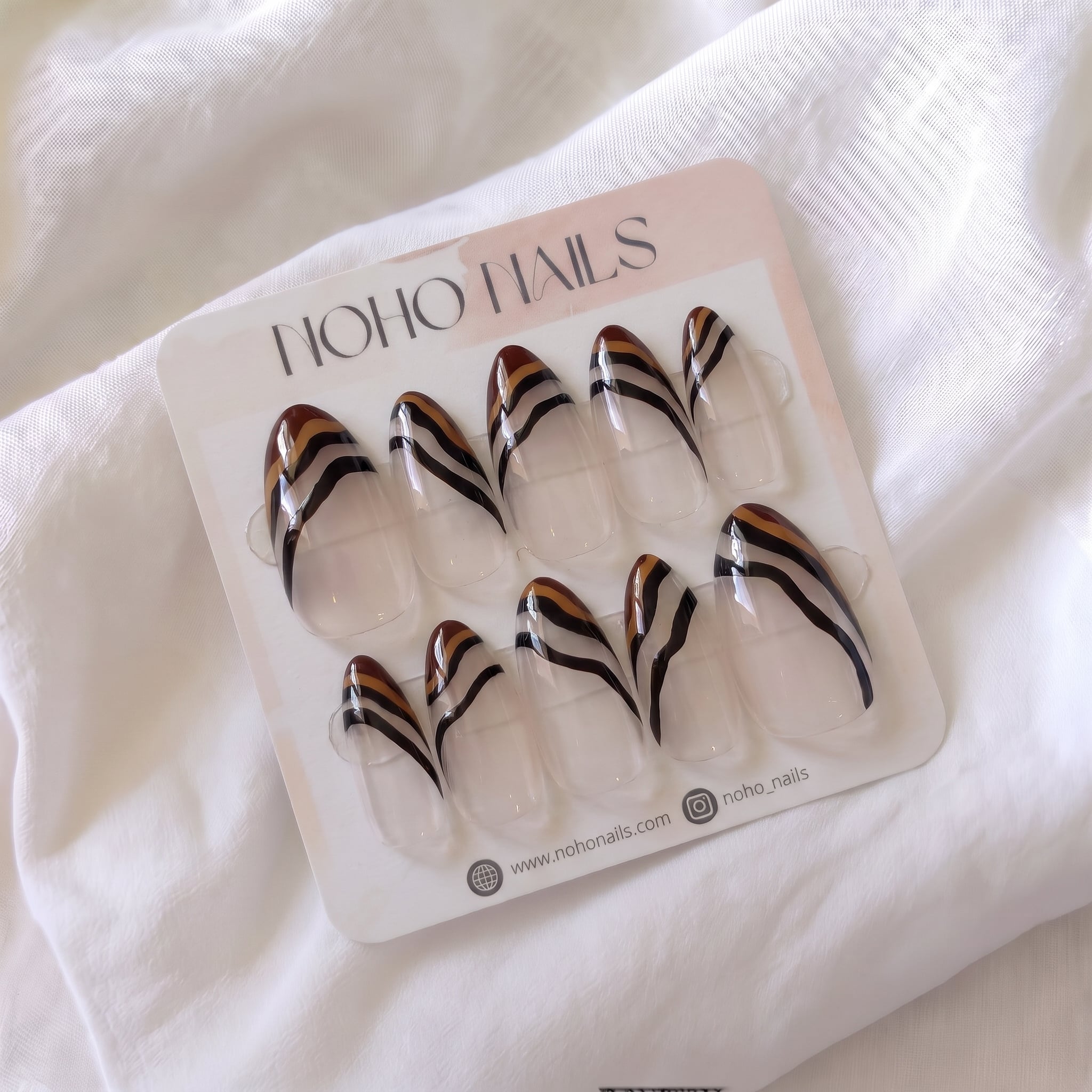 Ombre Portée press-on nails by Noho Nails: long almond nails with a sheer clear base, chocolate-brown tips and wavy caramel and black stripes on a Noho Nails display card