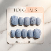 Slate Relief press-on nails on a Noho Nails display card lying on white marble: ten matte dusty blue-gray nails with a raised hand-sculpted texture