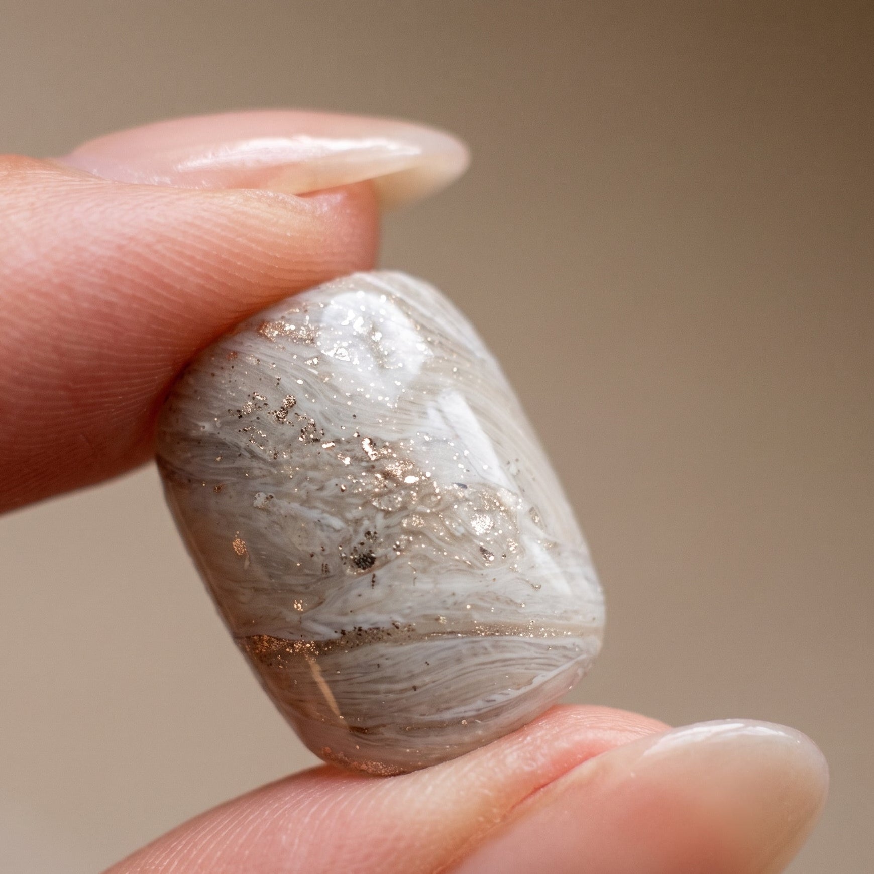 Fingers holding a single Sediment No. 9 press-on nail, showing greige and white marbled swirls with gold flecks and a glossy faceted surface