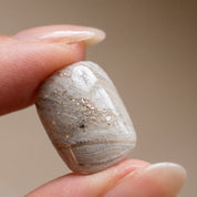 Fingers holding a single Sediment No. 9 press-on nail, showing greige and white marbled swirls with gold flecks and a glossy faceted surface