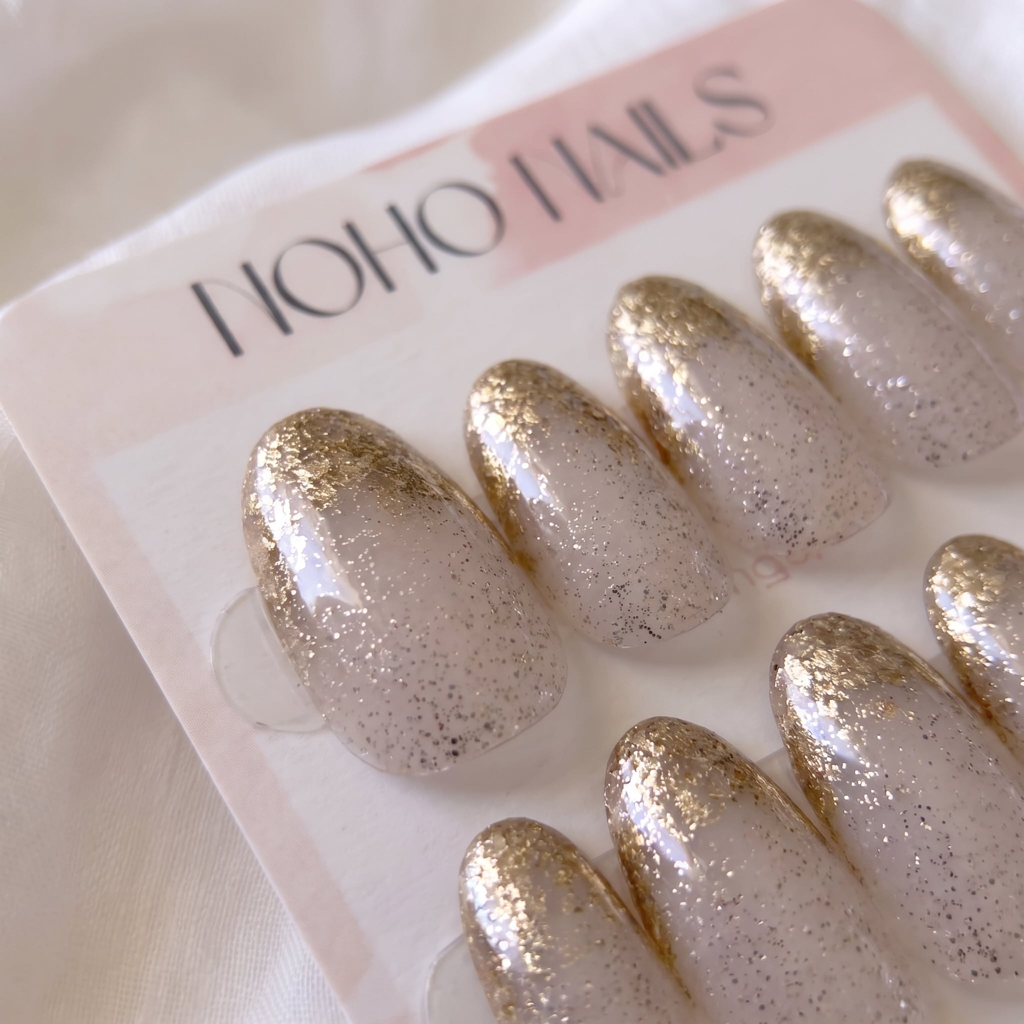Close-up of Golden Glaze press-on nails by Noho Nails: sheer nude oval nails with speckled glitter and gold glitter tips