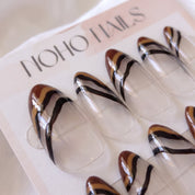 Close-up of Ombre Portée press-on nails by Noho Nails: long almond nails with a sheer clear base, chocolate-brown tips and wavy caramel and black stripes