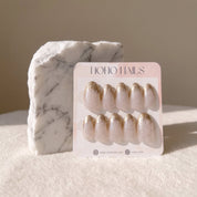 Golden Glaze press-on nails on a Noho Nails display card propped against a white marble stone