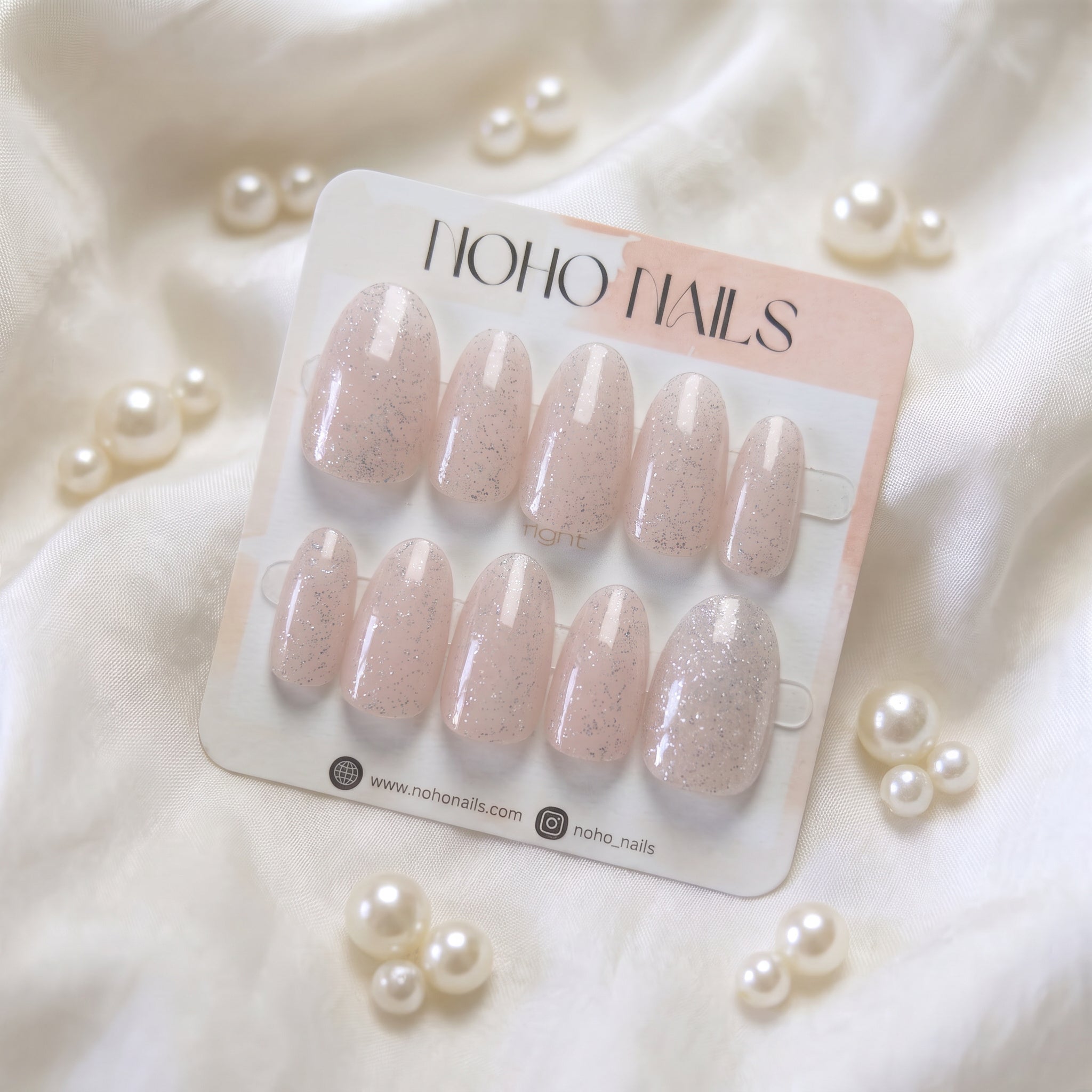 A set of ten HOHO NAILS press-on false nails in a soft blush pink glitter shade, displayed on their white and pale pink packaging card, styled on a white satin fabric with decorative pearls.