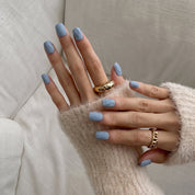 Hands wearing Slate Relief press-on nails in a short squoval shape, matte dusty blue-gray with a raised textured finish, with gold rings and a cream knit sweater